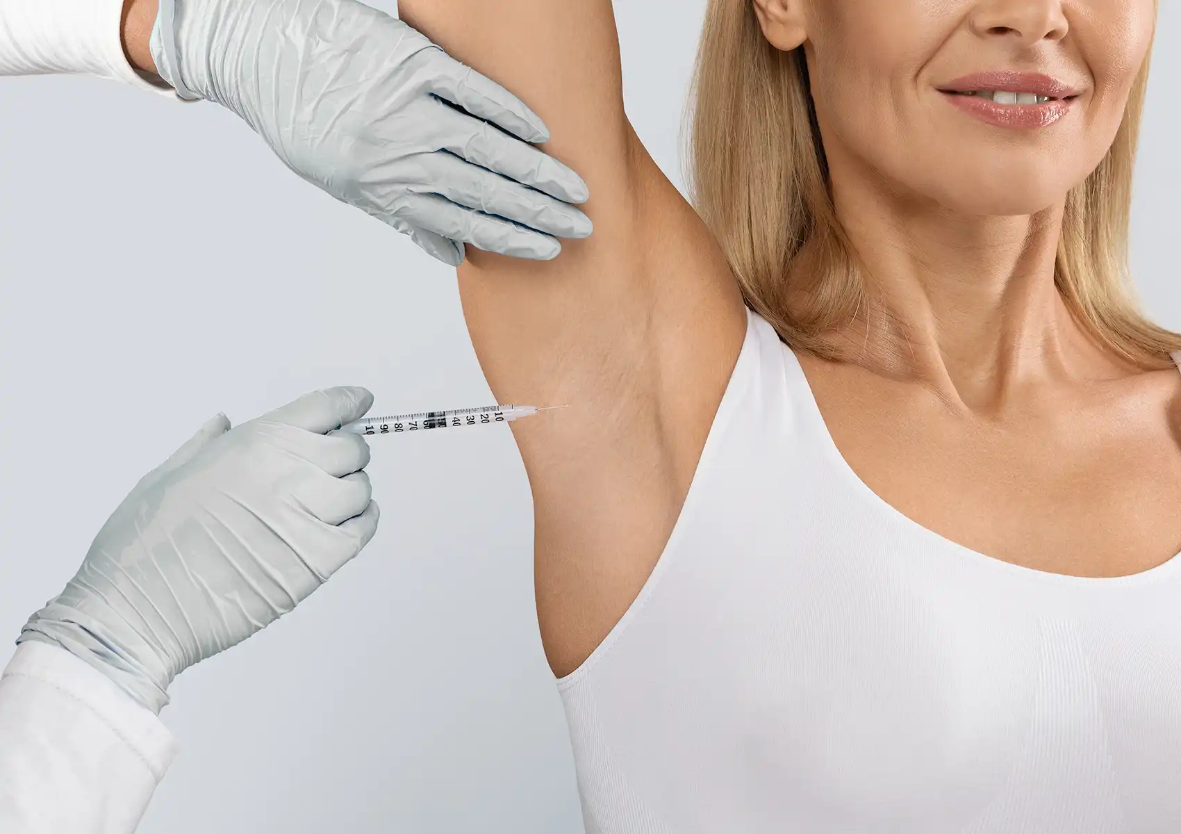 A gloved medical professional administers a syringe injection into the underarm of a smiling woman wearing a white tank top, demonstrating the procedure for botox for excessive sweating.