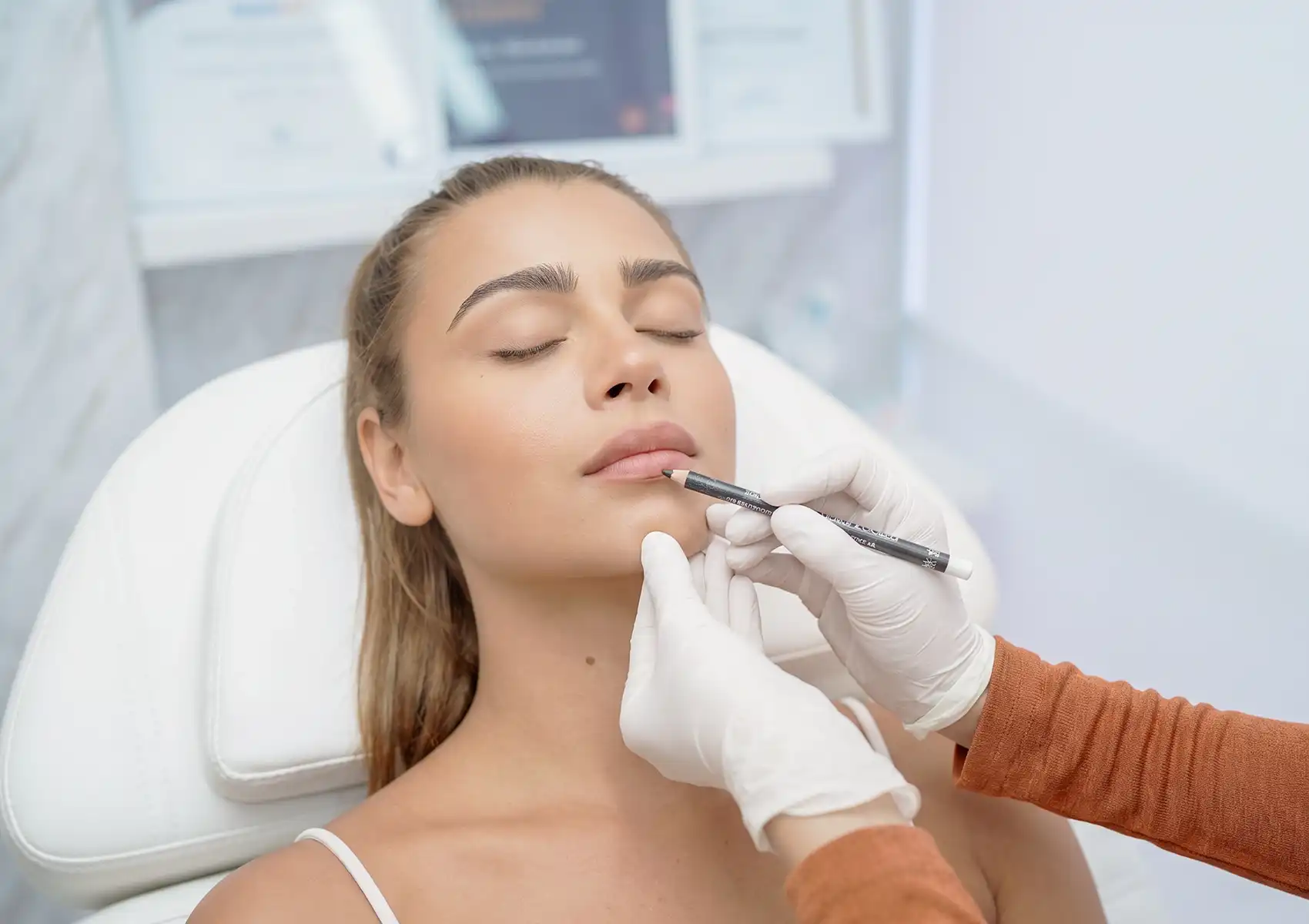 A practitioner in white gloves uses a white makeup pencil to carefully mark the lips of a female patient reclining with her eyes closed, preparing for a lip flip Bali procedure.