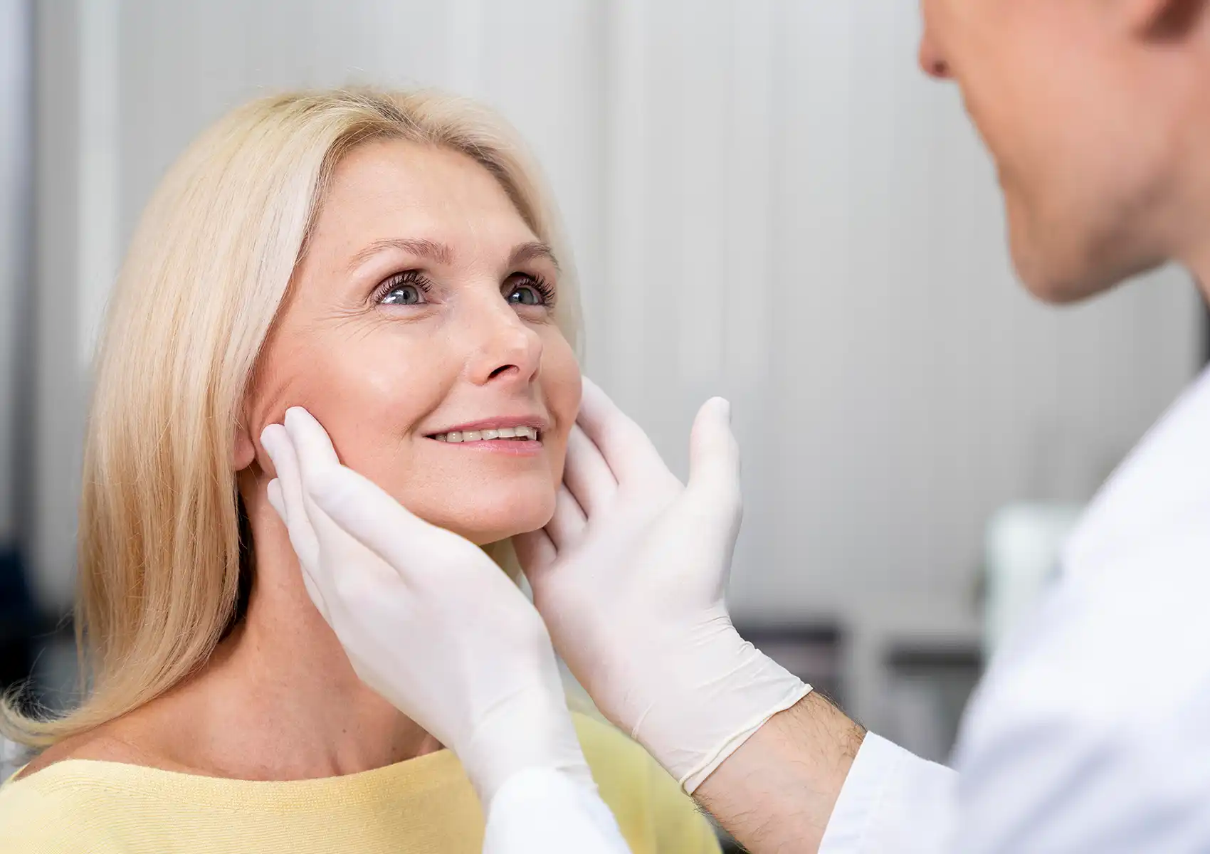 A gloved medical professional gently examines the face of a smiling, mature blonde woman during a consultation for anti-aging treatments at a doctor-led aesthetic clinic Bali