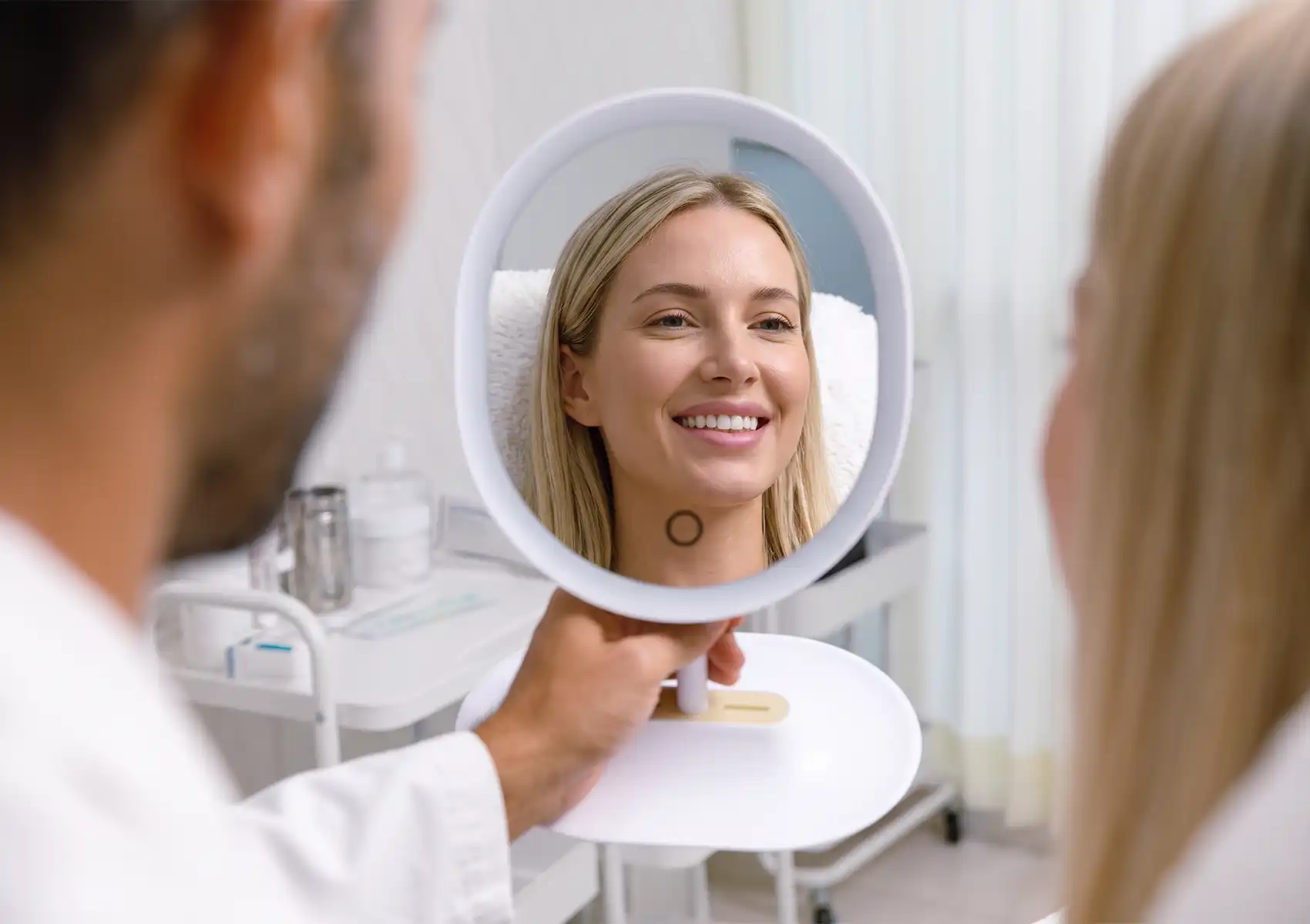 A smiling blonde woman looks into a brightly lit hand mirror held by a medical professional, evaluating her facial aesthetics during a consultation for a gummy smile treatment.