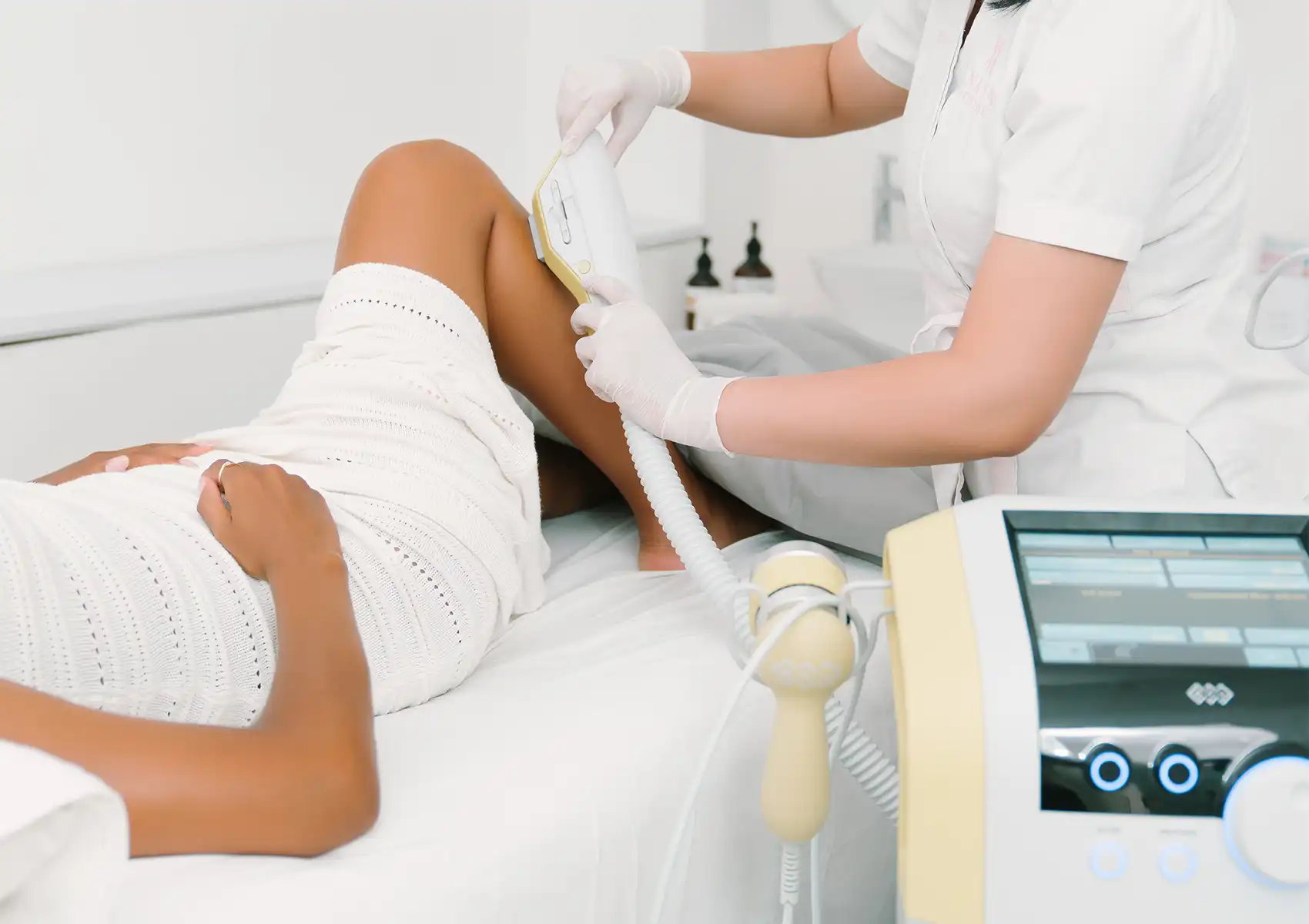 A practitioner wearing white gloves performs Laser hair removal on a female patient's lower leg using a modern aesthetic machine in a bright, clean clinic setting.