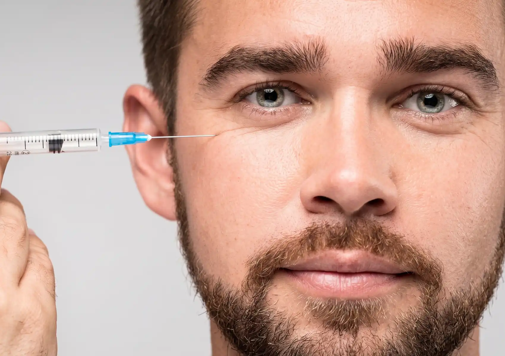 A close-up portrait of a man looking directly at the camera while a gloved hand holds a syringe near the outer corner of his eye, representing anti-aging treatments like botox for men Bali.