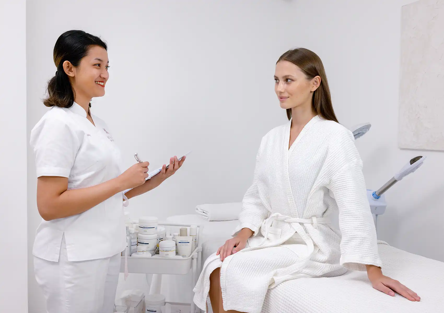 A smiling practitioner in a white uniform holding a clipboard while consulting with a client in a white robe seated on a treatment bed at a beauty clinic bali.