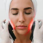 A woman with her eyes closed receiving red LED light therapy on her cheeks from a gloved professional at a beauty clinic bali.
