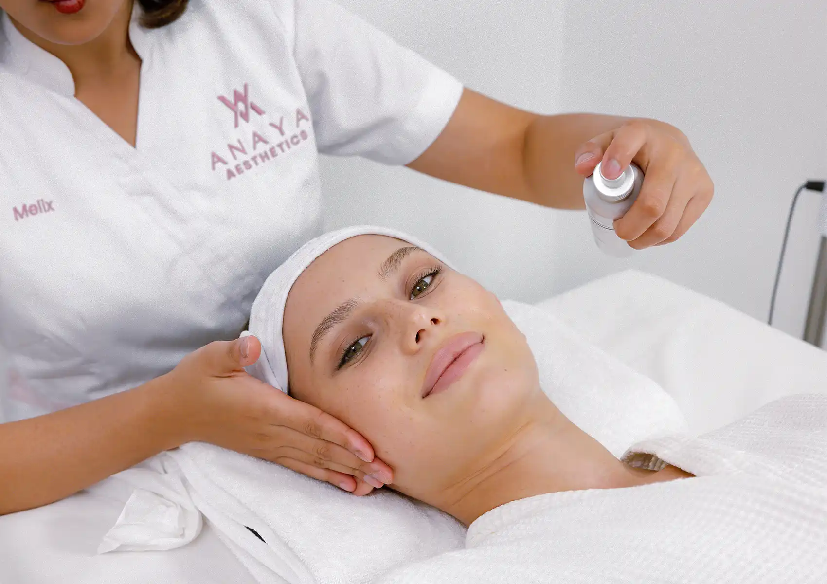 A skincare specialist at a beauty clinic bali spraying a refreshing mist onto a relaxed woman's face during a pre-wedding skin preparation treatment.