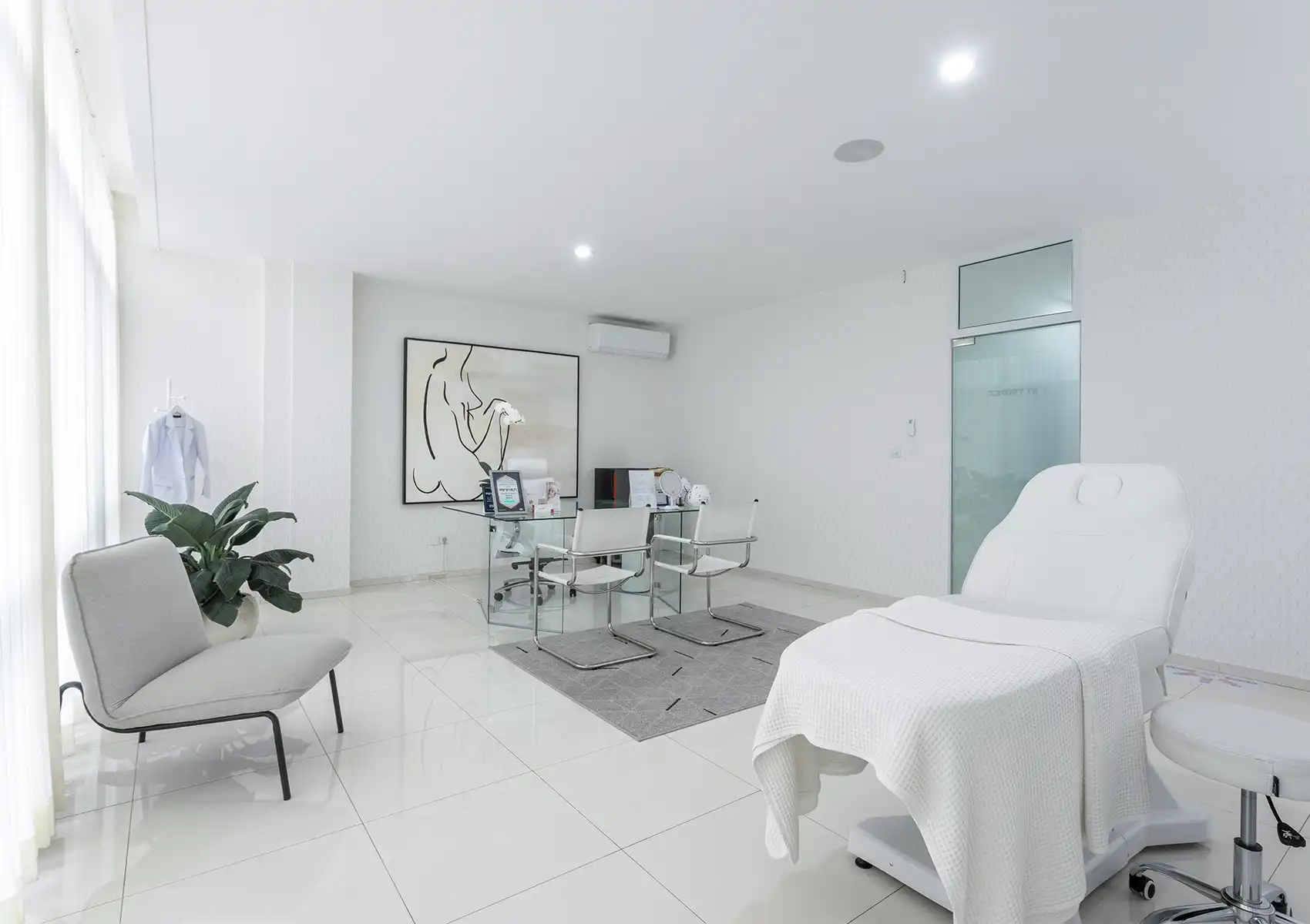 A bright, modern consultation and treatment room featuring a glass desk, chairs, and a white aesthetic bed at a beauty clinic bali.