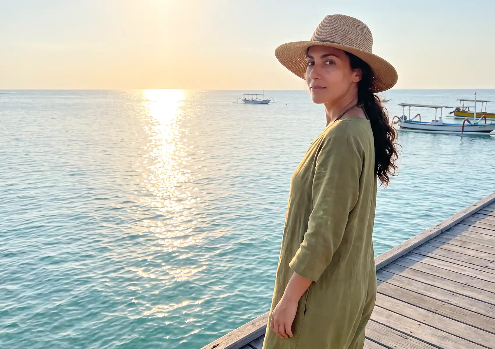 A woman wearing a sun hat on a wooden pier by the ocean, illustrating sun exposure that often prompts skin treatments at a beauty clinic bali.