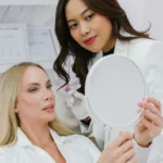 A doctor at Anaya Aesthetics in Kerobokan performing a facial assessment on a patient holding a mirror before a hyaluronic acid dermal filler treatment.