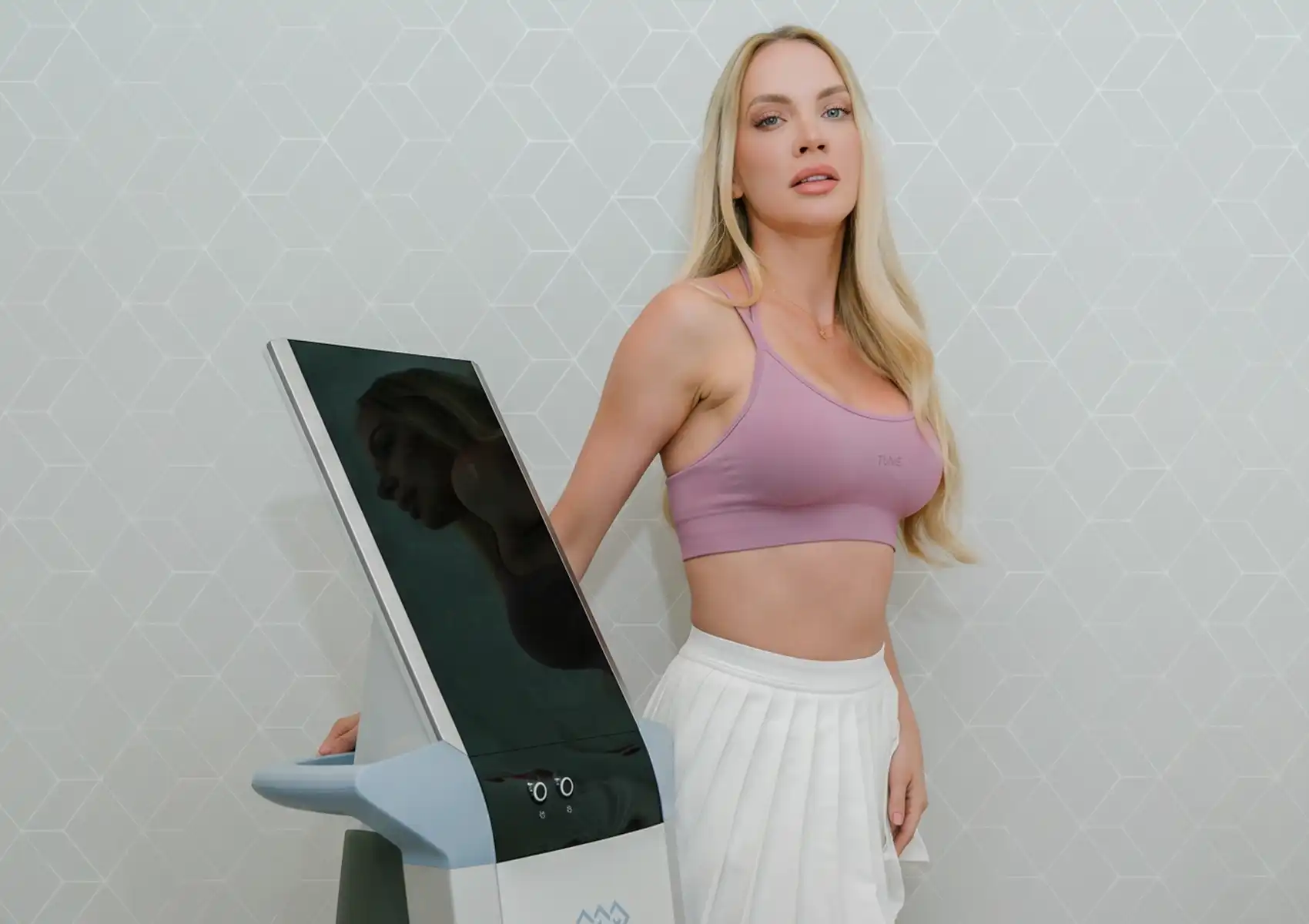 A blonde woman in a pink sports bra and white skirt standing confidently next to a modern Emsculpt body contouring machine at a beauty clinic bali.