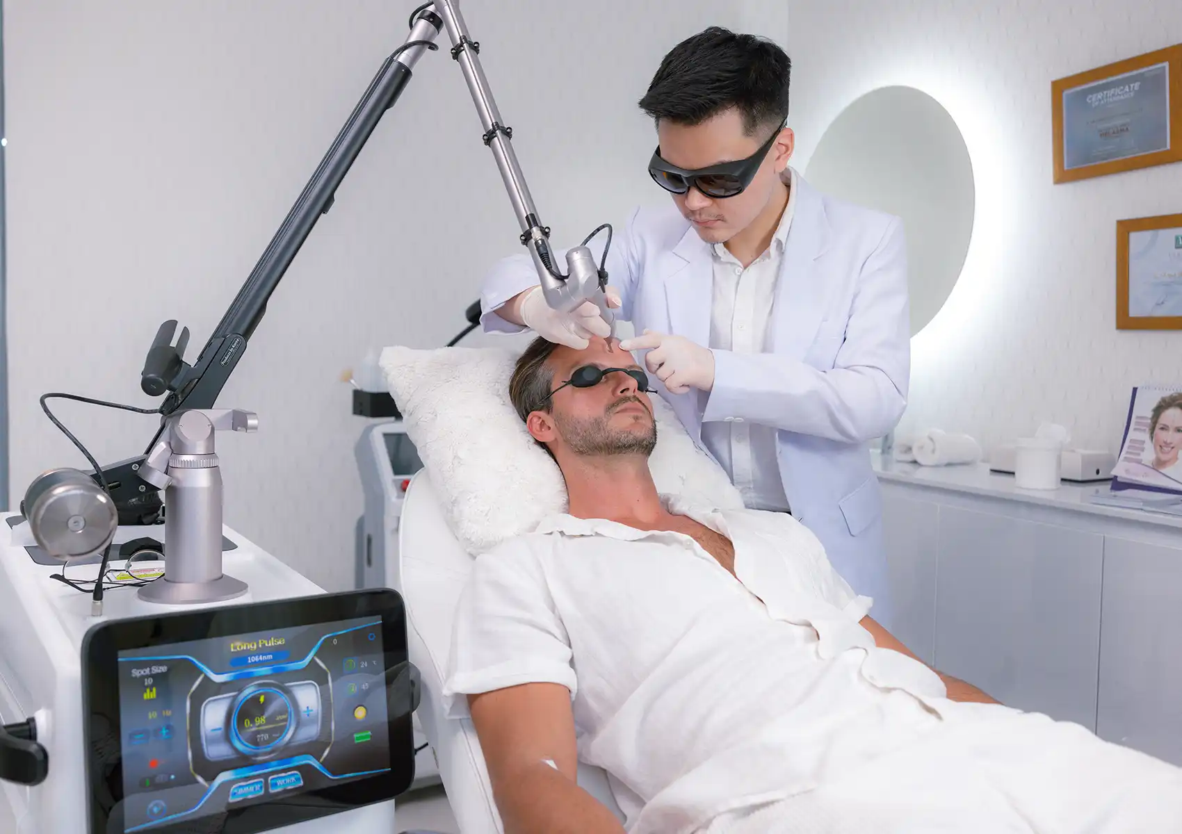 A male practitioner wearing a white coat and safety goggles carefully operates a large laser machine with a digital display to treat a patient's forehead area in a modern beauty clinic bali setting